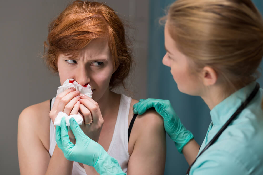 Foto de mulher ruiva com o nariz sangrando sendo atendida por uma médica para ilustrar conteúdo de sangramento nasal.